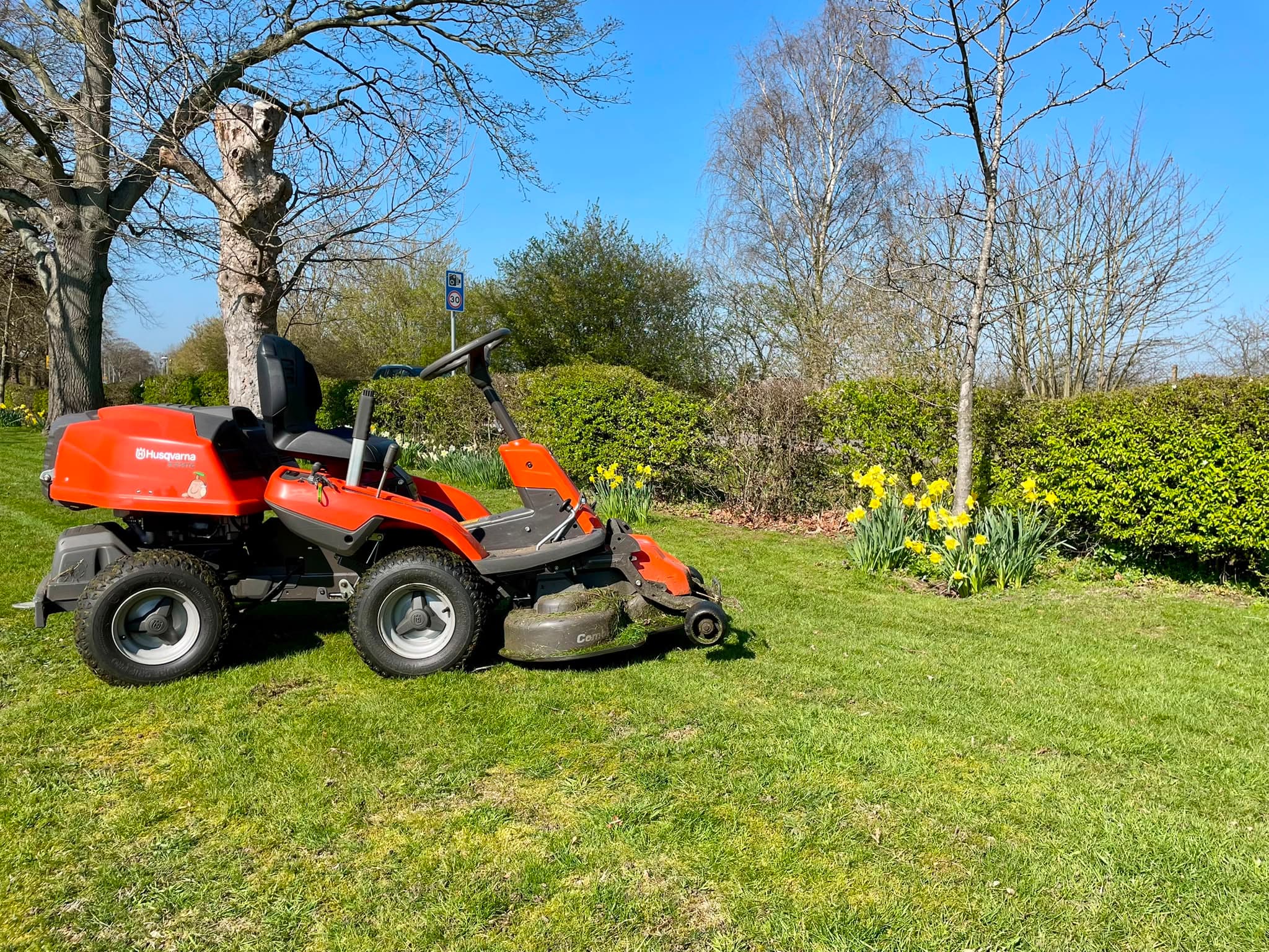 A ride-on mower parked on a large freshly cut lawn, with daffodils and a clipped hedge along the boundary