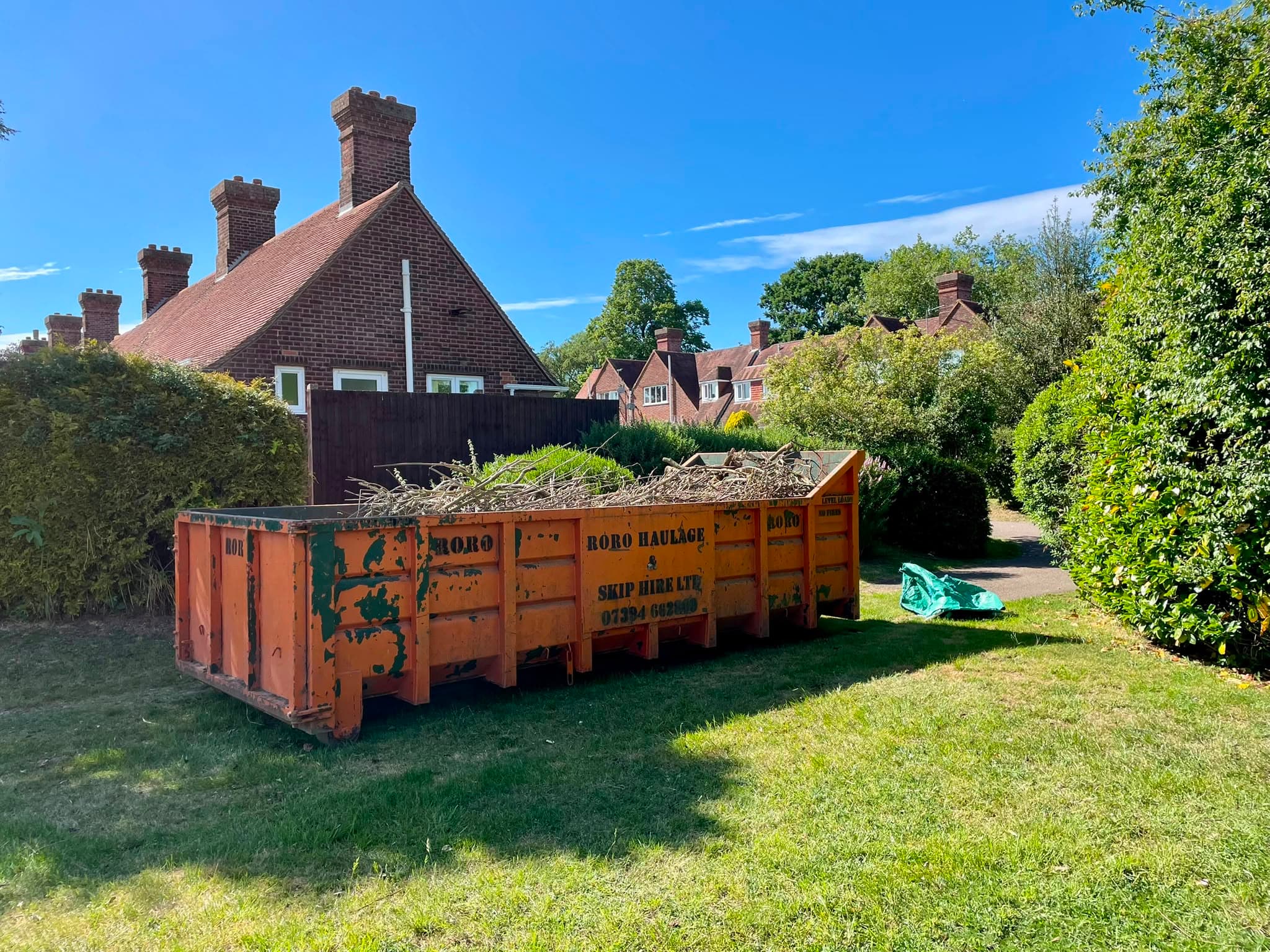 A large orange roll-on skip standing on a lawn, loaded with cut branches and hedge waste ready to be taken away
